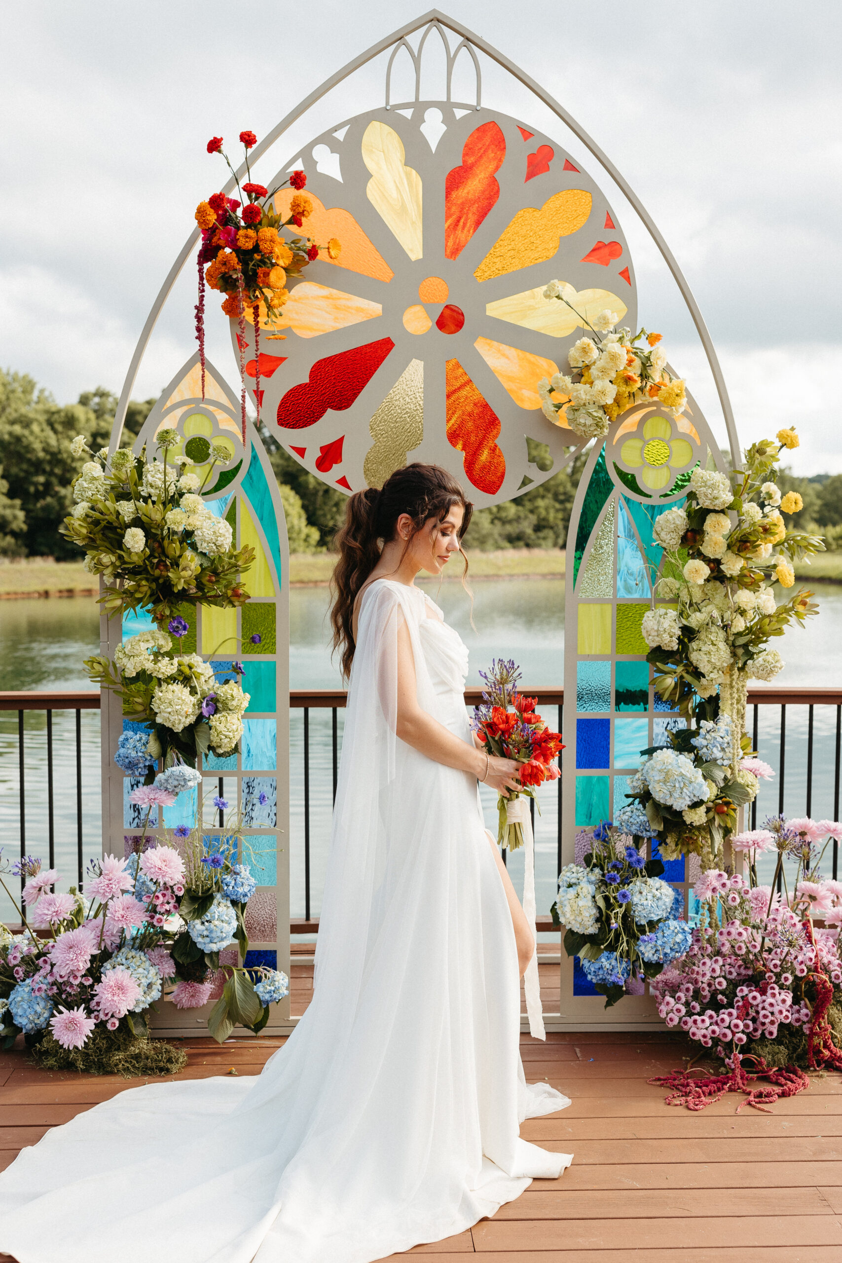 Bride in modern white gown standing in front of a vibrant stained glass arch adorned with rainbow florals at the water's edge.
