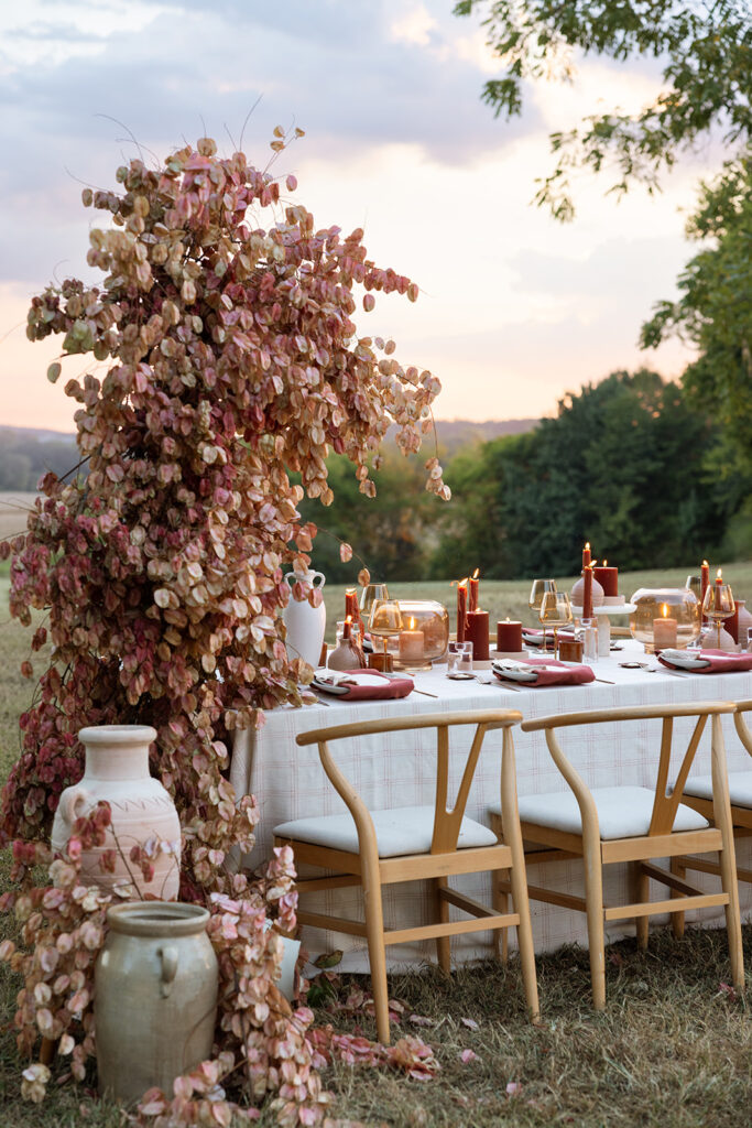 Fall wedding tablescape under a Chinese flame tree installation with terracotta vessels, rust candles, blush napkins, and wishbone chairs styled in an open field.