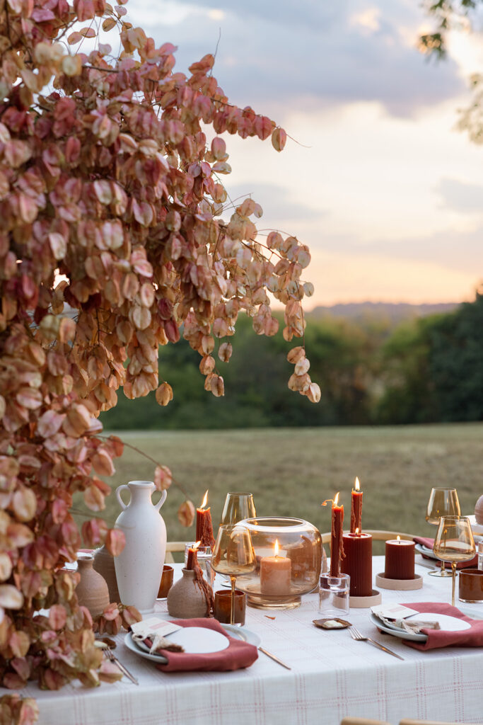 Fall wedding table styled under Chinese flame tree branches with terracotta vessels, amber glassware, rust candles, and blush napkins at sunset.