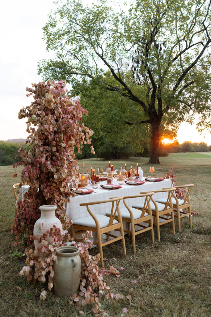 Wide fall wedding tablescape under a tree at sunset with Chinese flame tree branches, terracotta vessels, rust candles, blush linens, and wishbone chairs.
