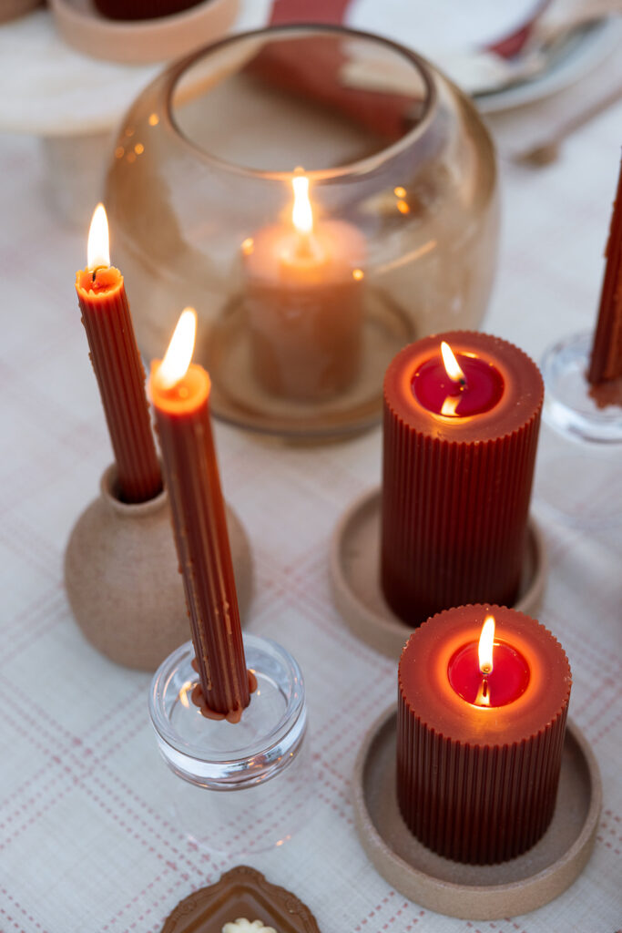 Close-up of ribbed rust candles glowing in terracotta and glass holders on a fall wedding table styled with a blush linen grid cloth.