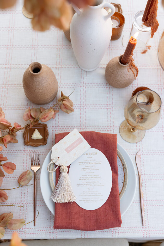 Fall wedding place setting with rust linen napkin, round menu card, tassel detail, amber glassware, and clay vessels styled with Chinese flame tree branches.