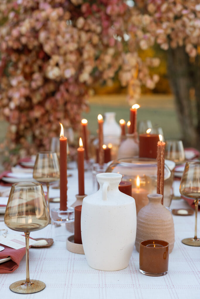 Editorial fall wedding tablescape detail with rust candles, terracotta vessels, white ceramic urns, and amber glassware styled with blush napkins.