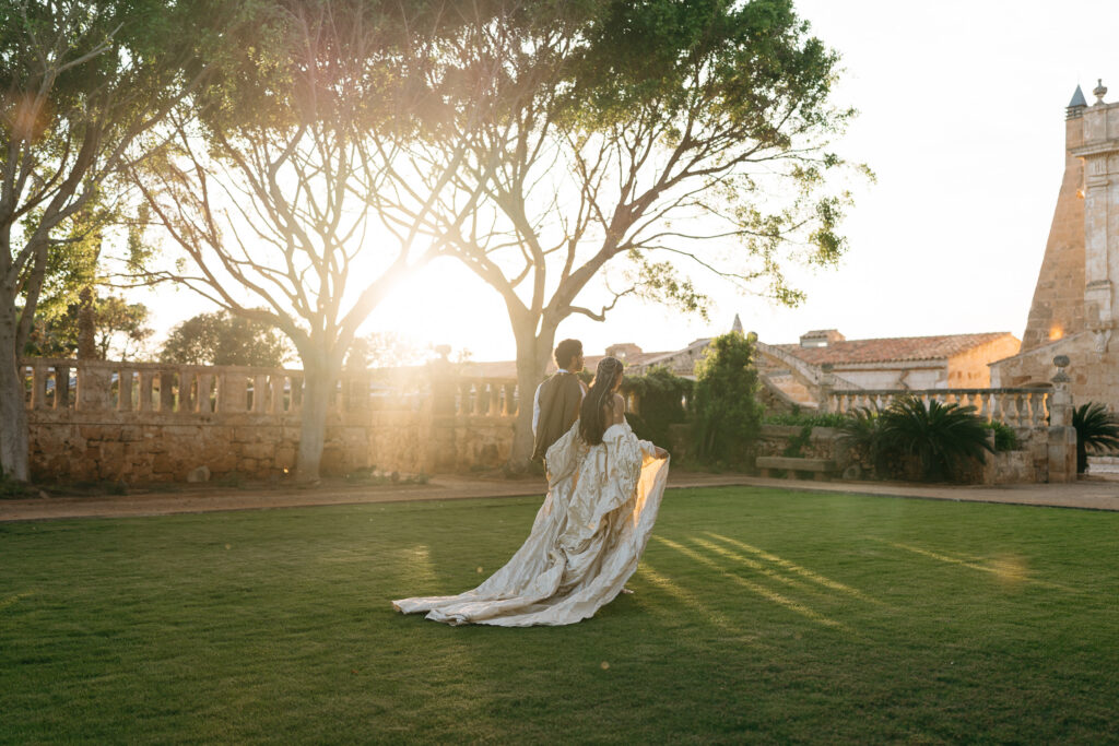 Bride and groom walk toward the sunset past a stone balustrade in the gardens at Vestige Son Vell, Menorca