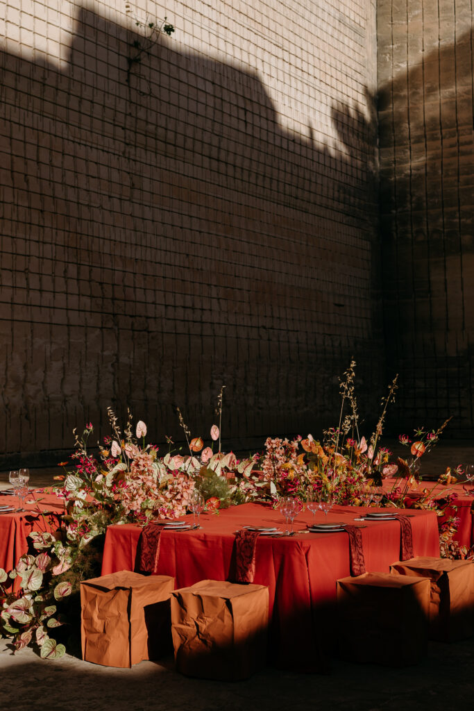 Terracotta wedding table with a low floral spine of anthuriums and wild grasses against the dark quarry wall, Lithica Quarry, Menorca — designed and produced by Fox & Quinn.