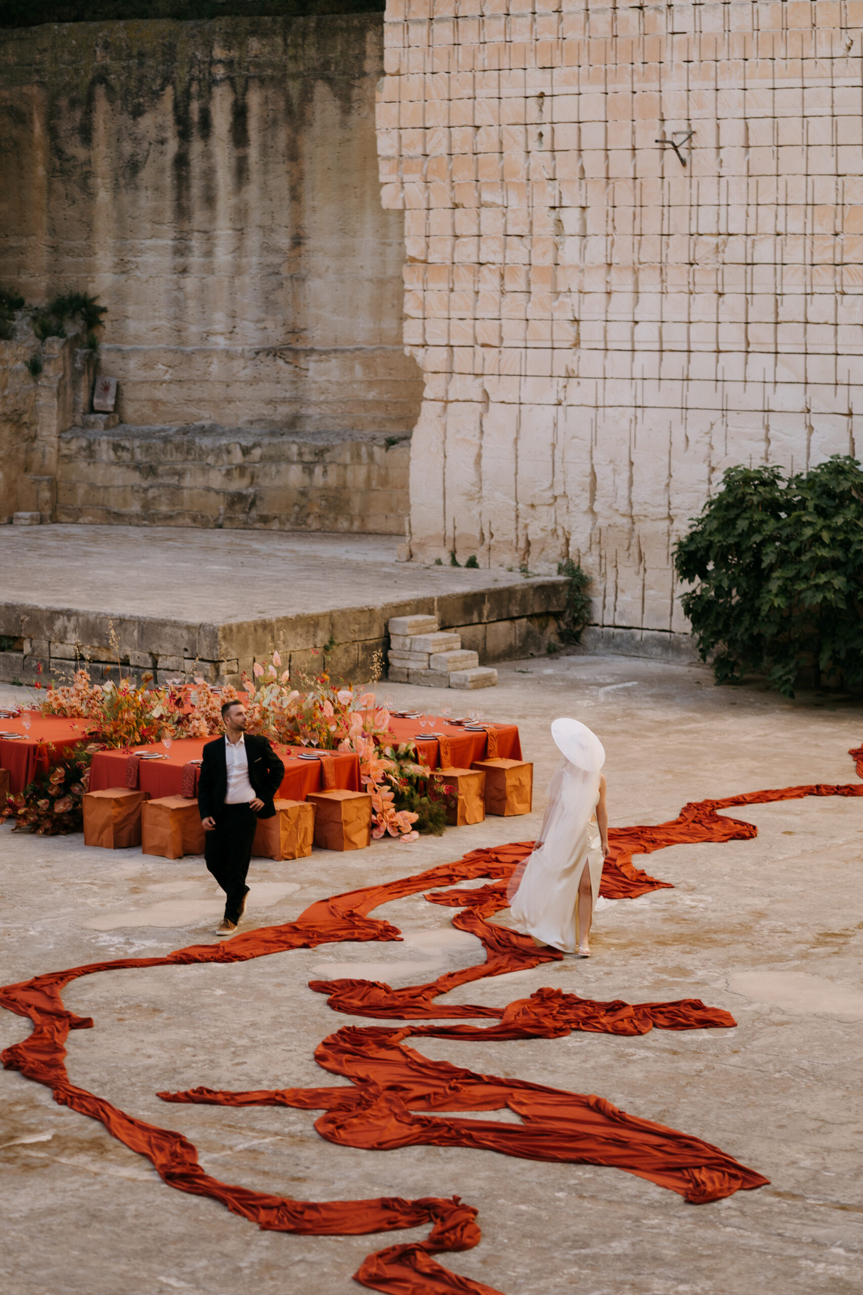 Bride walking a rust chiffon runner across the stone floor of the Amphitheatre quarry at Lithica, Menorca, toward terracotta dining tables — monochromatic destination wedding designed and produced by Fox & Quinn.
