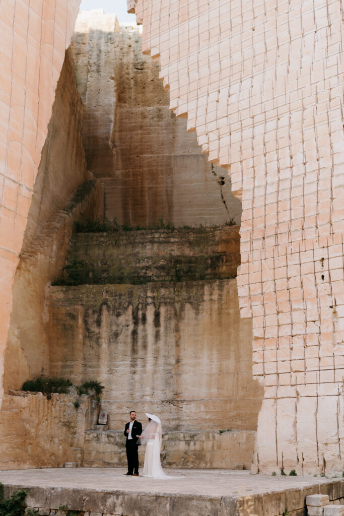 Bride and groom standing at the base of the towering stepped limestone walls of the Amphitheatre quarry at Lithica, Menorca — a dramatic destination wedding venue, designed and produced by Fox & Quinn.