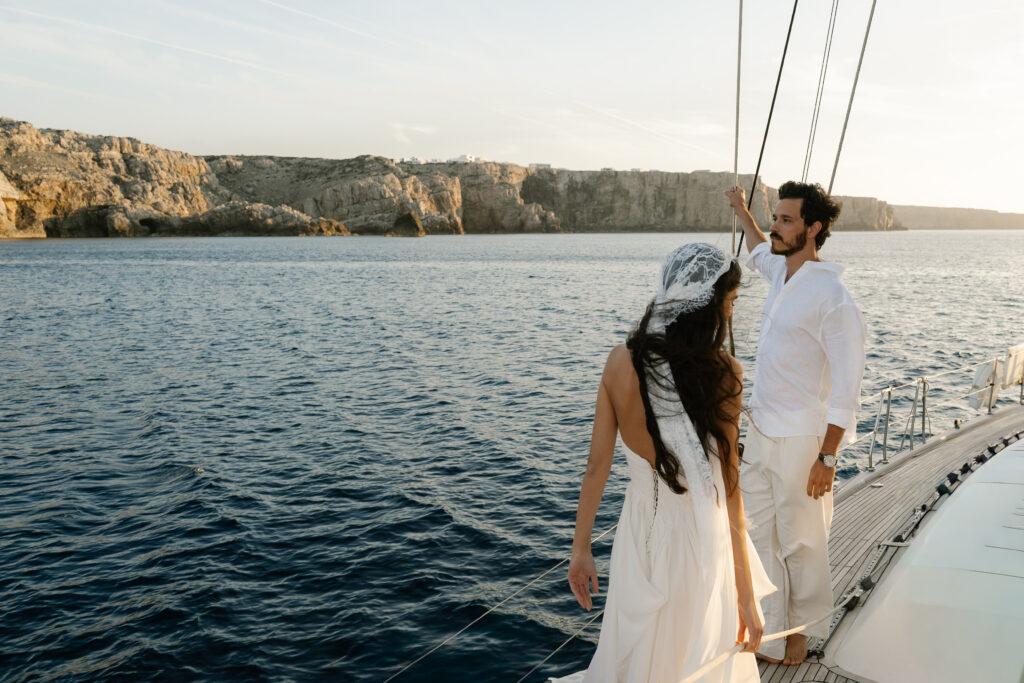 Bride in a lace veil aboard a boat passing the Menorca cliffs at golden hour
