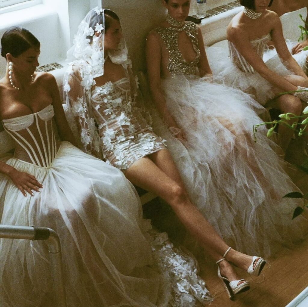 Group of Bronx and Banco models backstage at NYFW wearing corseted tulle and beaded bridal gowns in soft lighting.