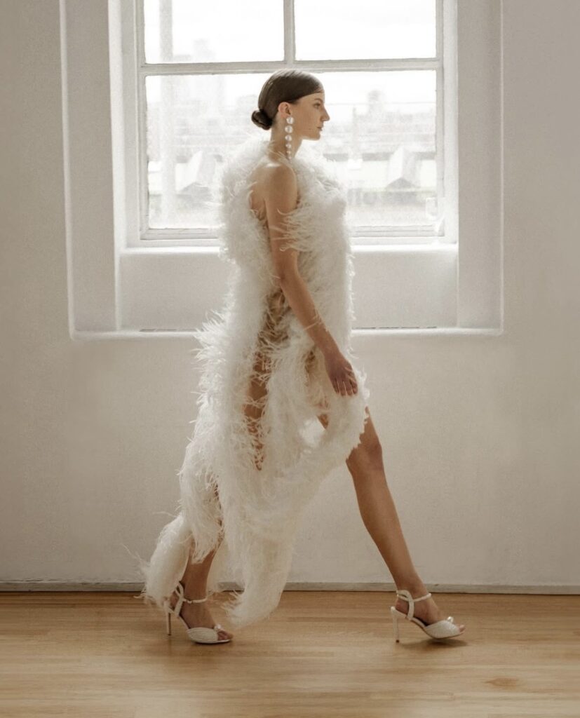 Model walking in a feathered bridal gown by Bronx and Banco, with pearl drop earrings and stiletto heels at NYBFW SS’25.