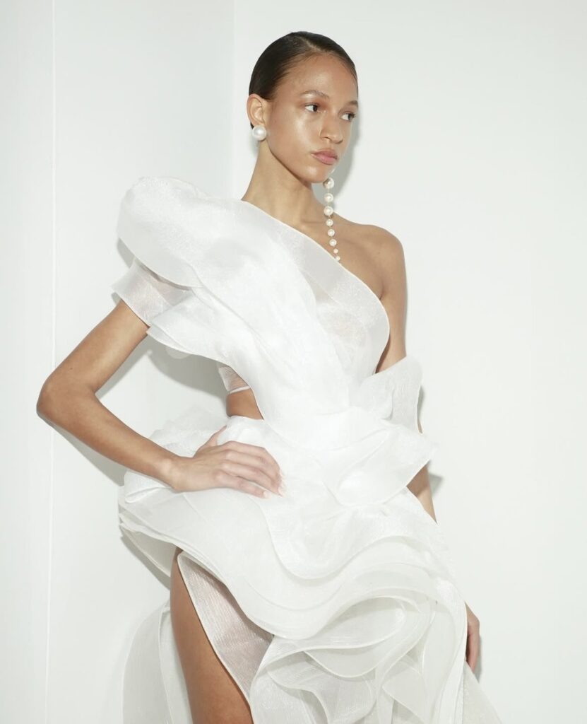 Close-up of a model wearing a sculptural one-shoulder white gown with pearl drop earrings and slicked-back hair.