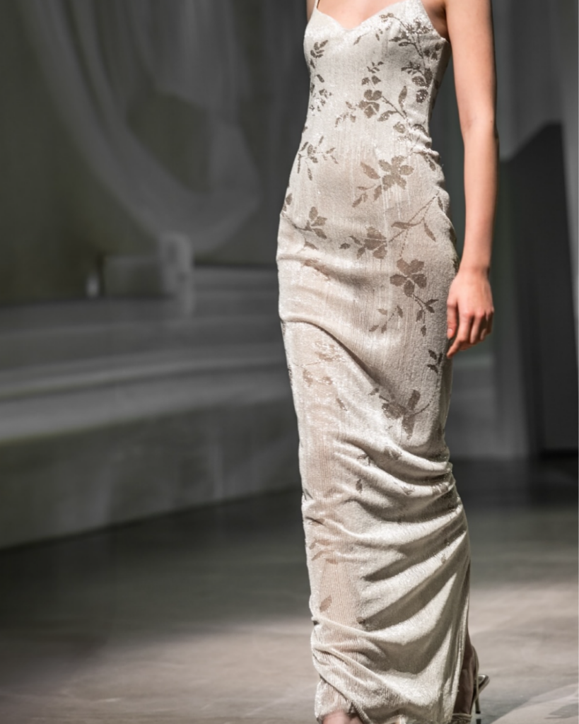 Model walking in a floor-length beaded ivory gown with botanical motifs and soft draping at the hem from EnAURA Bridal.