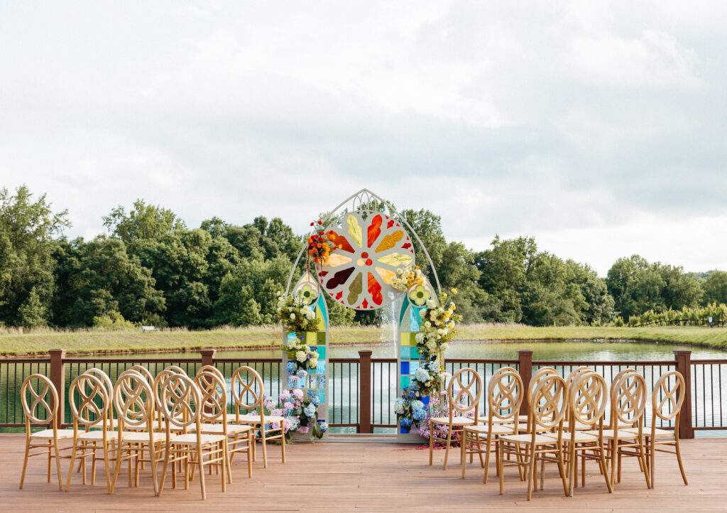 Outdoor wedding ceremony setup on a wooden deck overlooking the lake at Montaluce Winery. A custom arched structure mimics stained glass windows with bold colors and floral accents, surrounded by wooden oval-back chairs facing the altar.