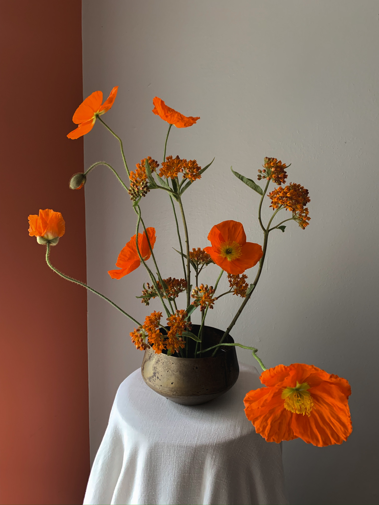 Sculptural floral arrangement of vivid orange poppies and clustered orange blooms in a bronze vessel, styled with minimalist elegance against a neutral backdrop.