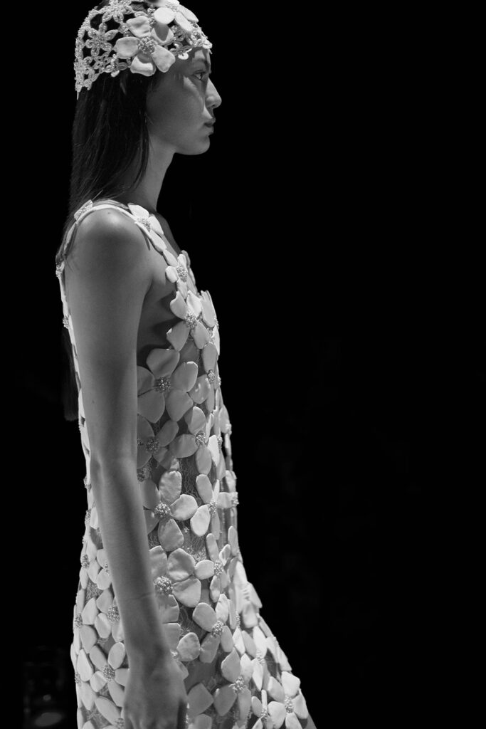 Profile view of a model wearing a sculptural gown with floral appliqués and a matching cap headpiece.