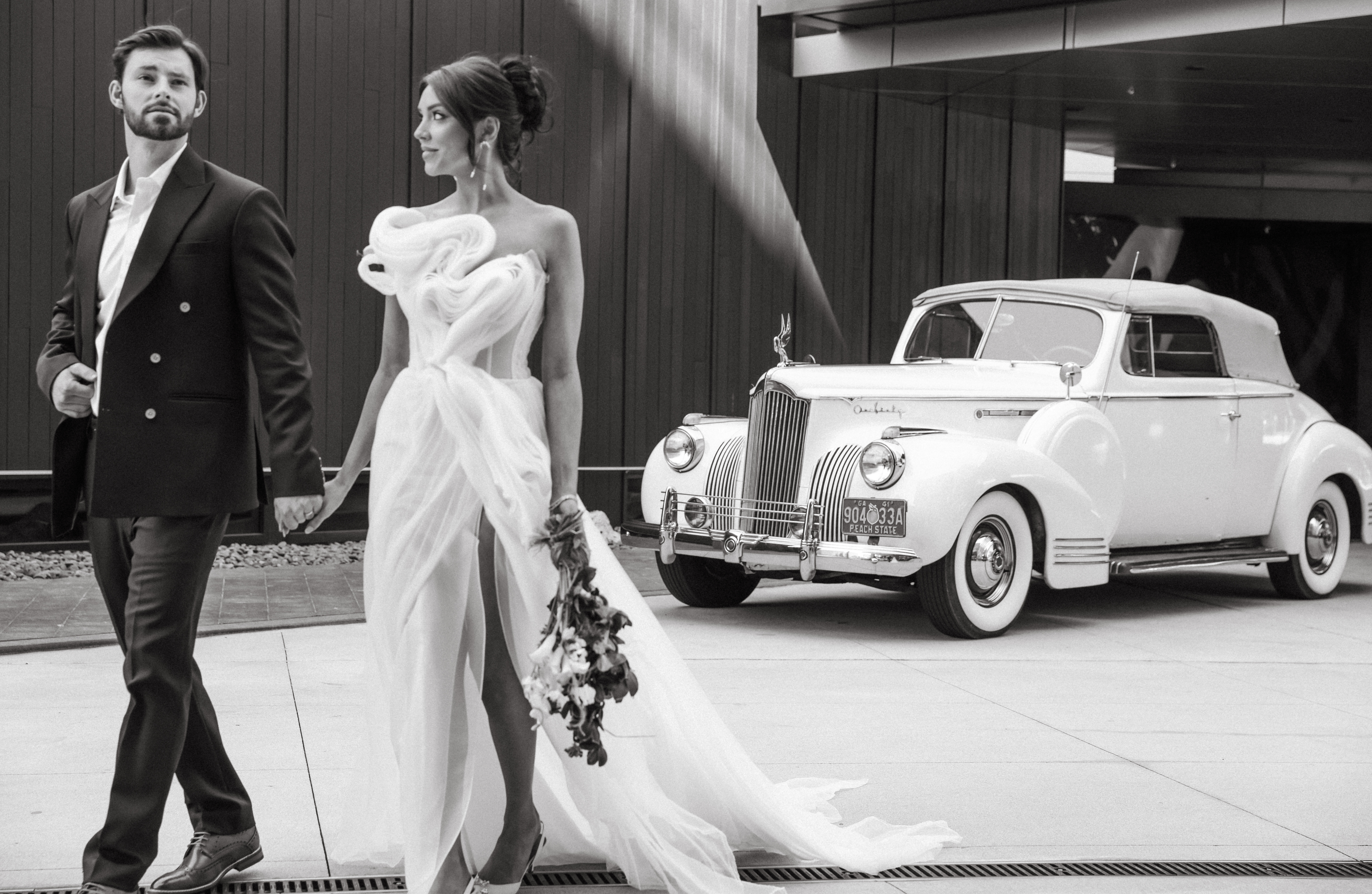 Black and white editorial wedding portrait of bride and groom walking hand-in-hand in front of a vintage white convertible, with the bride in a sculptural gown and the groom in a double-breasted suit.