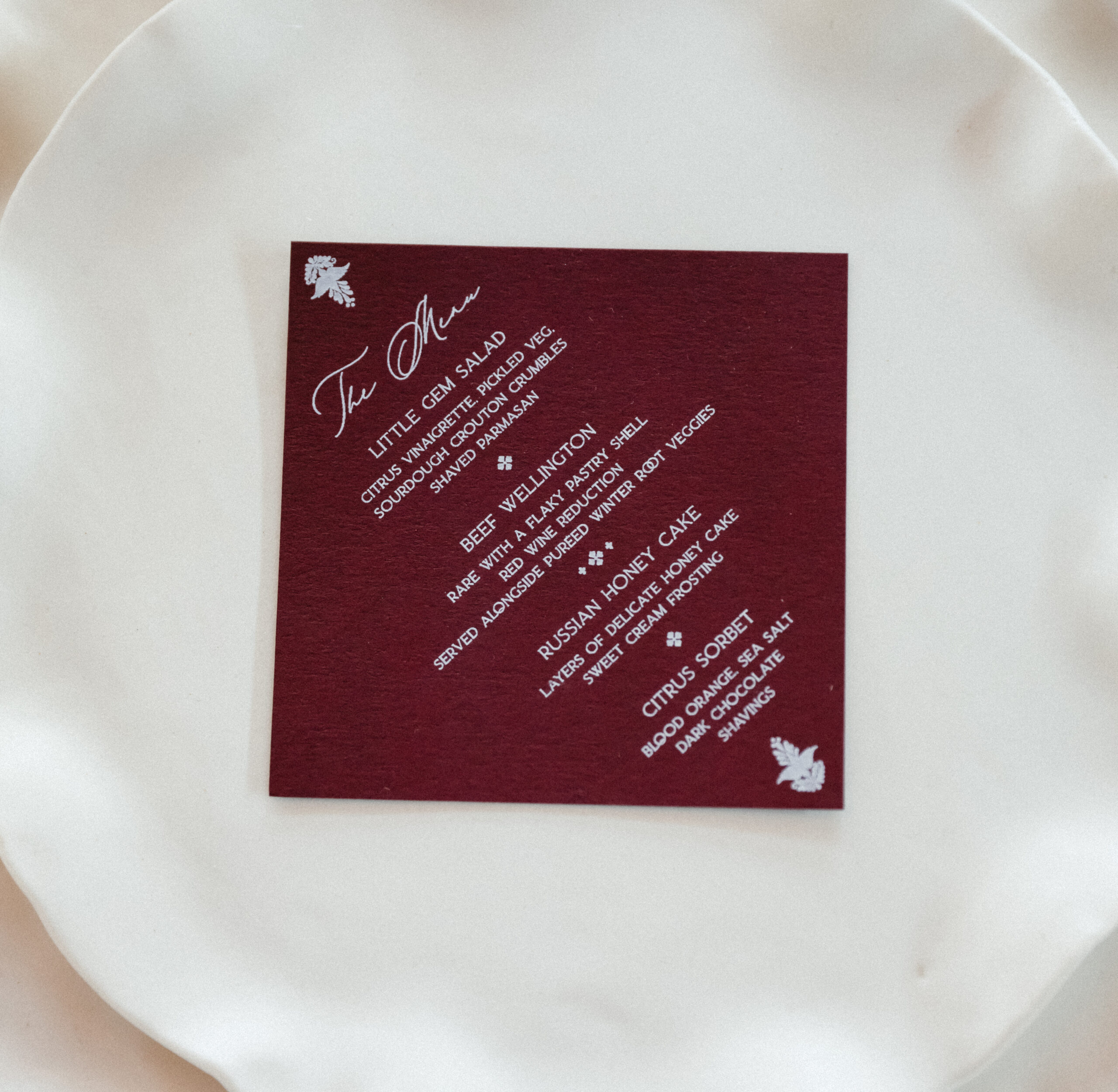 Burgundy modern wedding menu with white calligraphy-style text, styled diagonally on layered ivory ceramic dinnerware for a refined and minimalist tablescape.