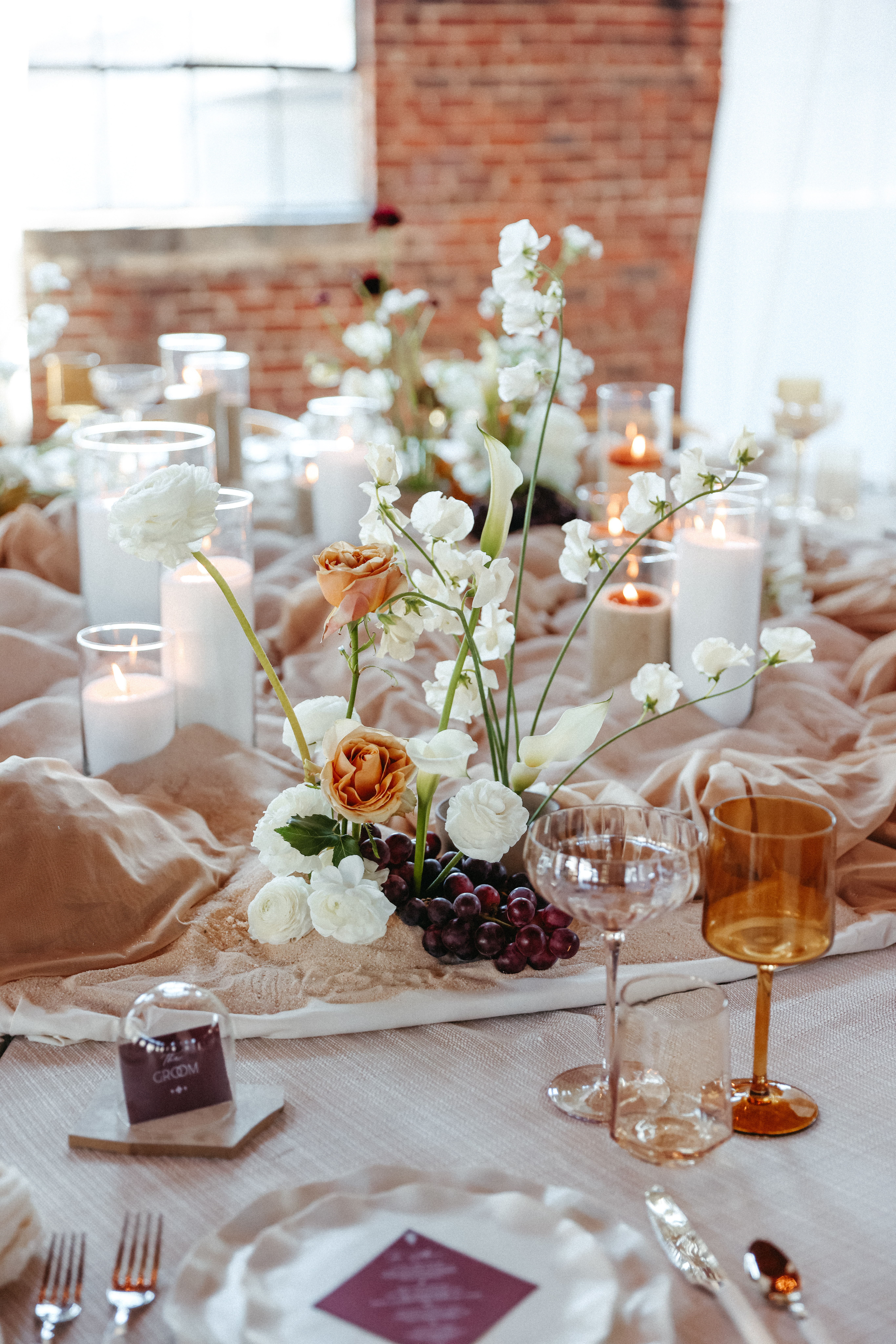Modern wedding tablescape featuring sculptural florals, neutral draping, pillar candles, amber-toned glassware, and a cluster of red grapes as an organic accent.