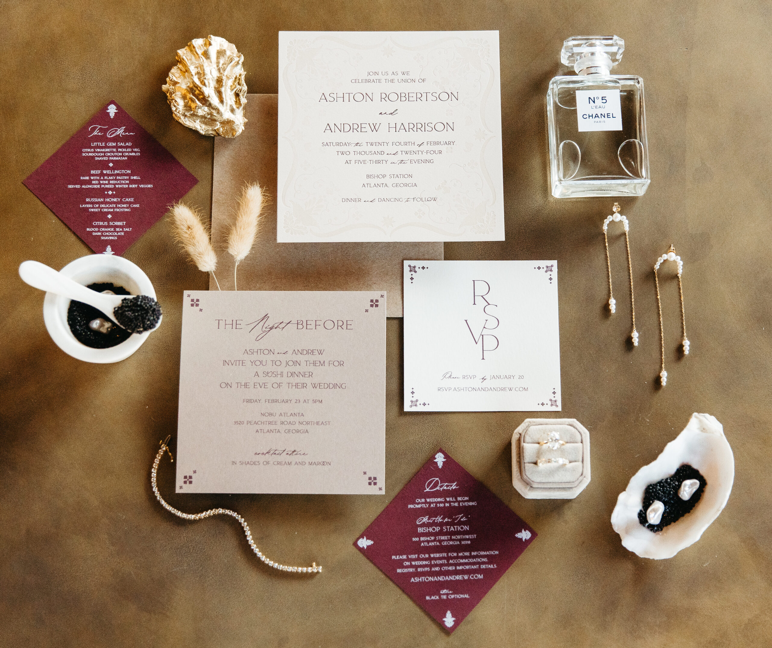 Flatlay of wedding invitation suite styled with burgundy and cream paper goods, black sea salt in a white shell dish, pearl earrings, a vintage-style ring, and Chanel No. 5 perfume bottle, all arranged on a warm brown velvet surface.