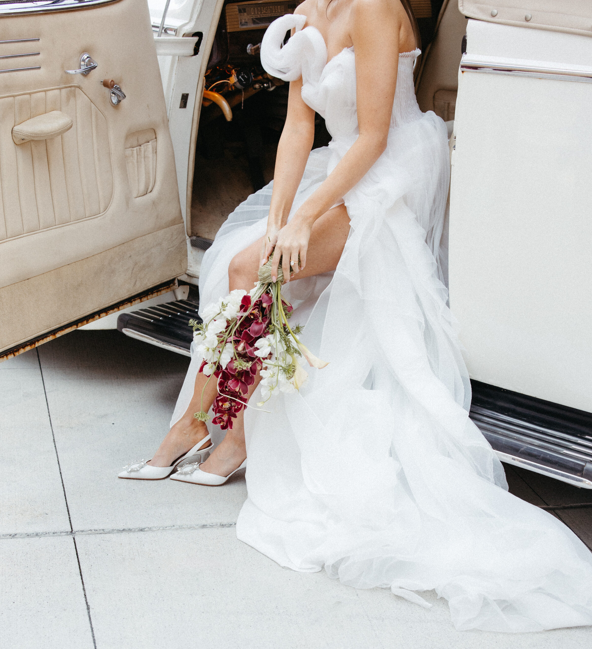 Close-up of bride sitting in a vintage convertible holding a sculptural bouquet with white and burgundy blooms, wearing a modern architectural gown.