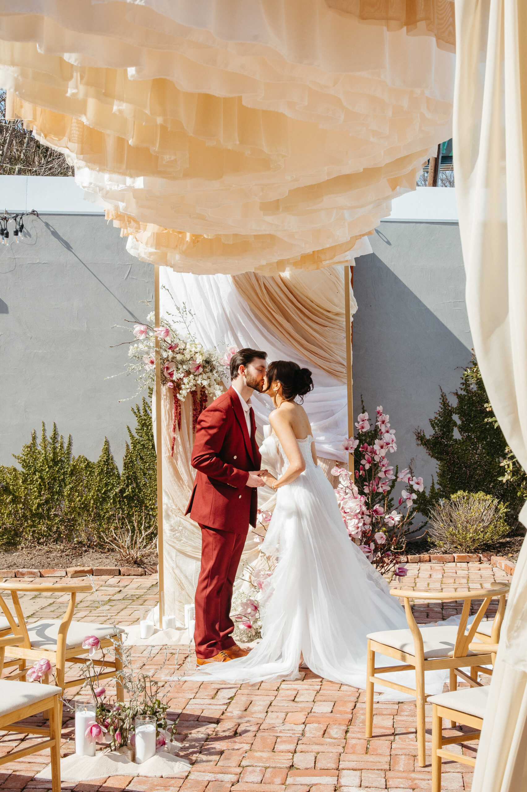 Bride and groom share a kiss under a layered ivory draped ceremony installation with cascading blush florals and golden frame, set against a stucco wall and spring greenery.