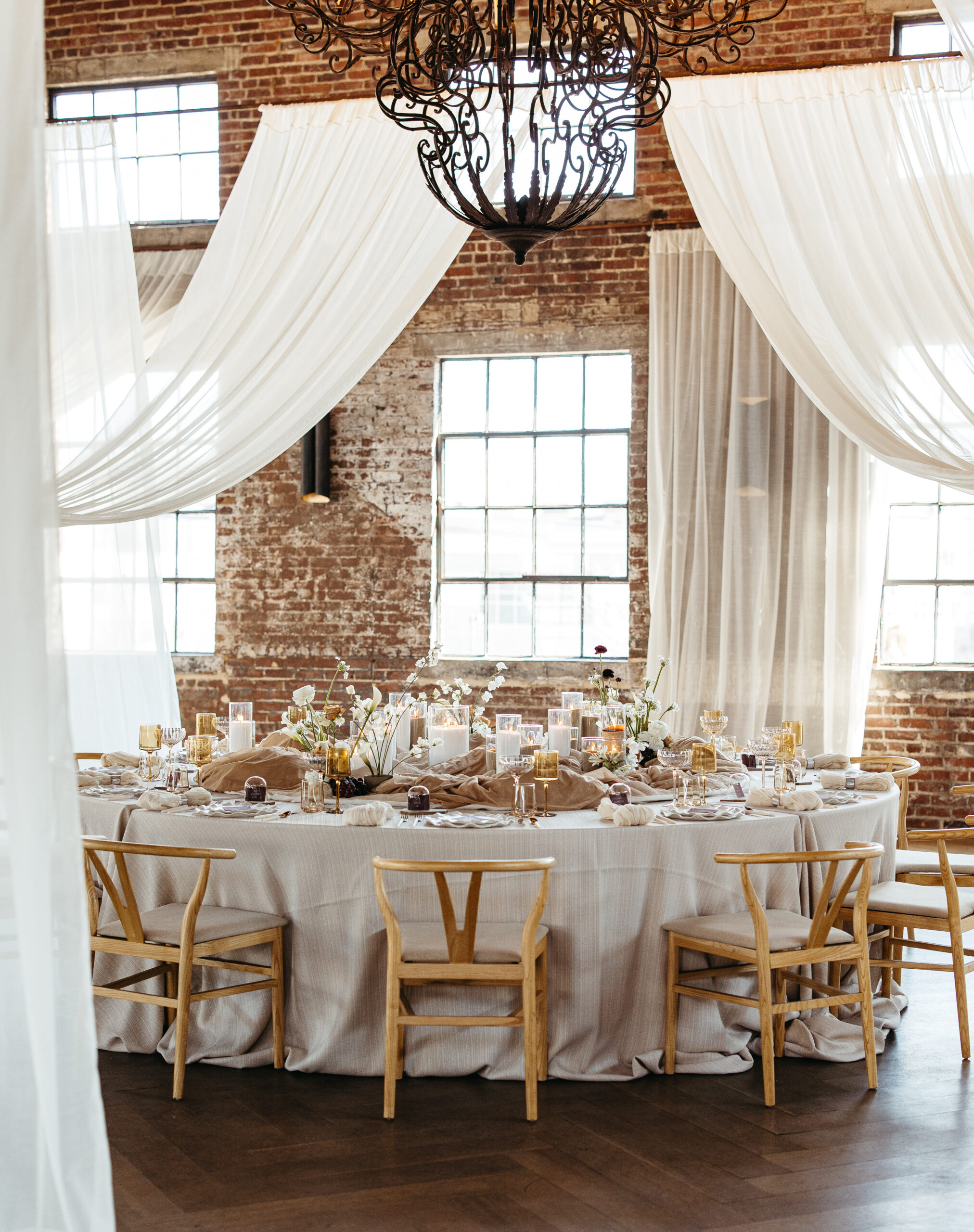 Round wedding reception table styled with neutral linens, modern ceramic vases, sculptural florals, and soft candlelight beneath sheer draping in an industrial loft with exposed brick walls and natural light.