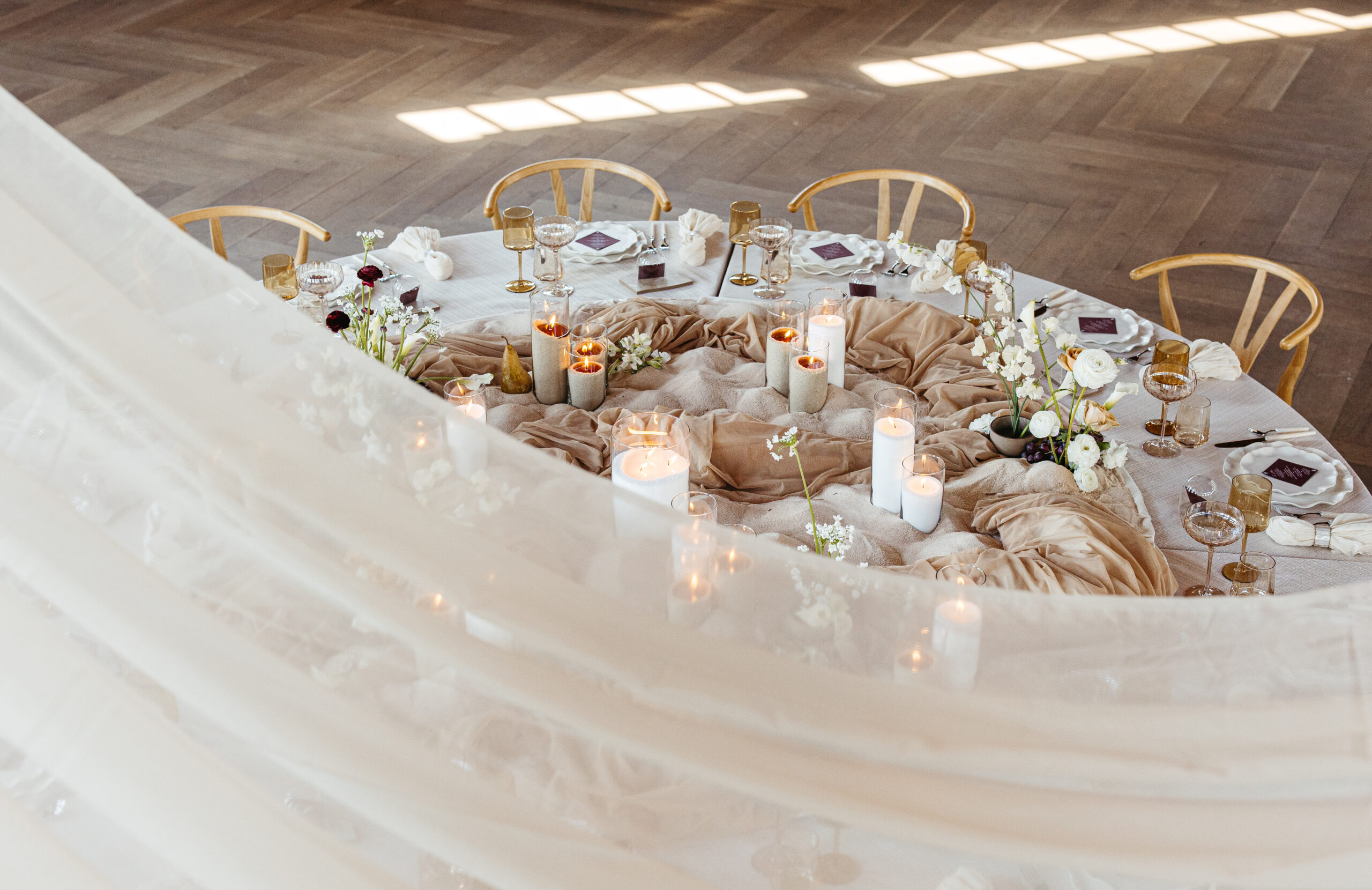 Overhead view of a modern round wedding table styled with sculptural tan draping, ivory pillar candles, minimalist floral arrangements, and neutral-toned place settings, softly framed by sheer fabric in a sunlit venue.