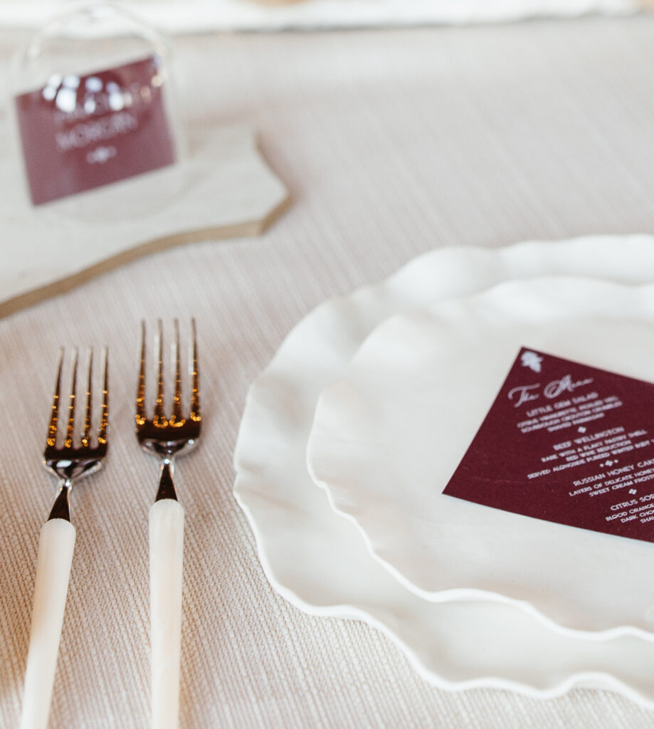 Elegant wedding place setting featuring layered white wavy-edged plates, cream-handled gold flatware, and a burgundy acrylic place card with modern typography set atop the plate on a soft neutral linen tablecloth.