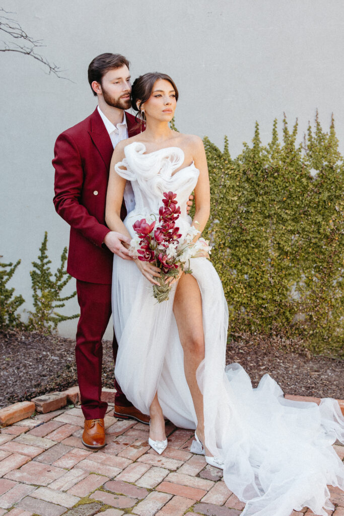 Bride and groom posing outdoors in front of a textured wall and greenery; the groom wears a burgundy suit, while the bride stands confidently in a sculptural, asymmetrical white gown holding a bouquet of red and blush blooms.