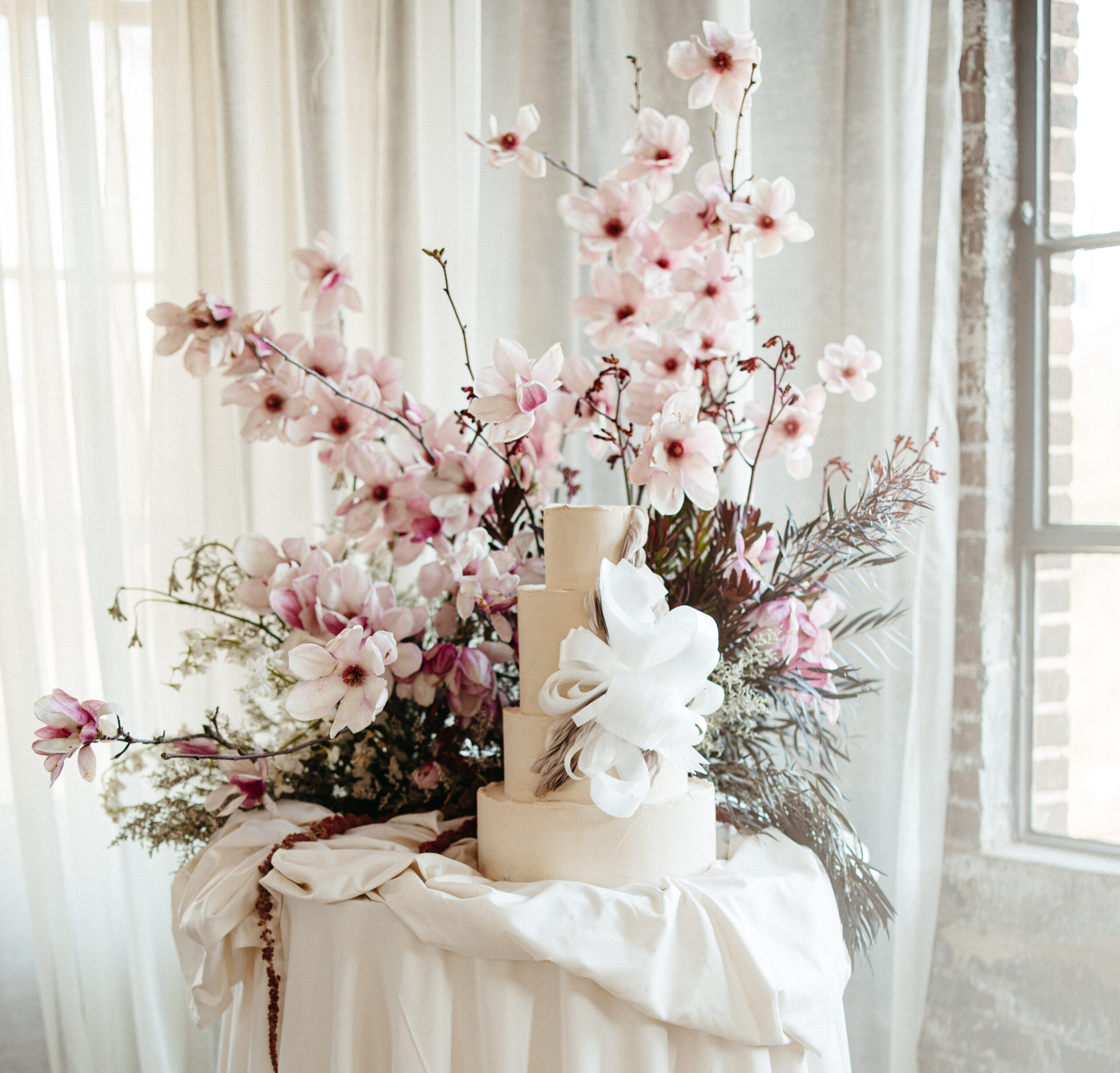 Modern ivory wedding cake with sculptural sugar detail, styled against cascading pink magnolia blooms and soft drapery in a sunlit space.