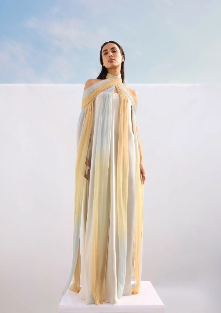 Billowing, off-the-shoulder chiffon gown in soft sky blue and muted peach hues, crisscrossing at the neckline and cascading into delicate pleats. Light as air and effortlessly celestial.