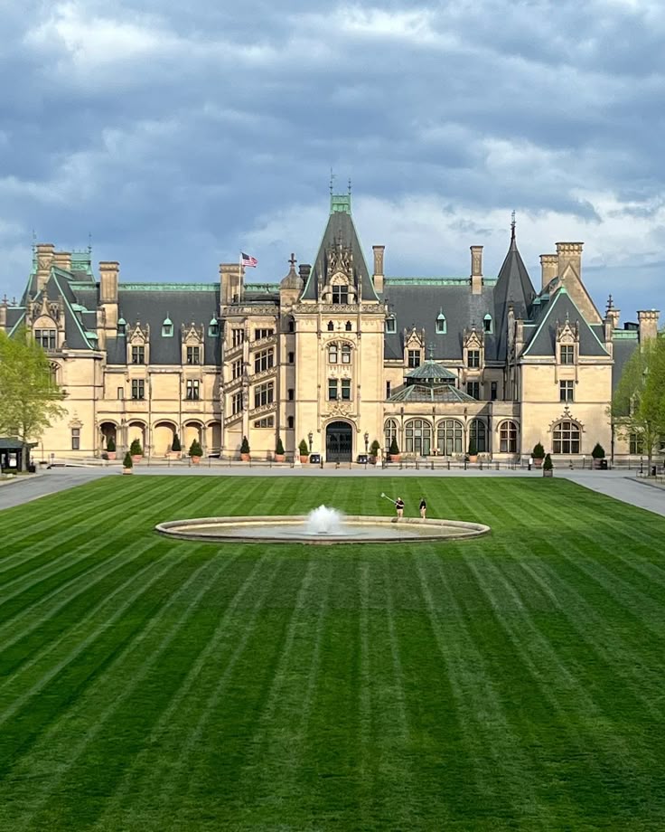 The grand facade of the Biltmore Estate rises beyond a perfectly manicured lawn, with a central fountain and two people walking near it under a moody sky.