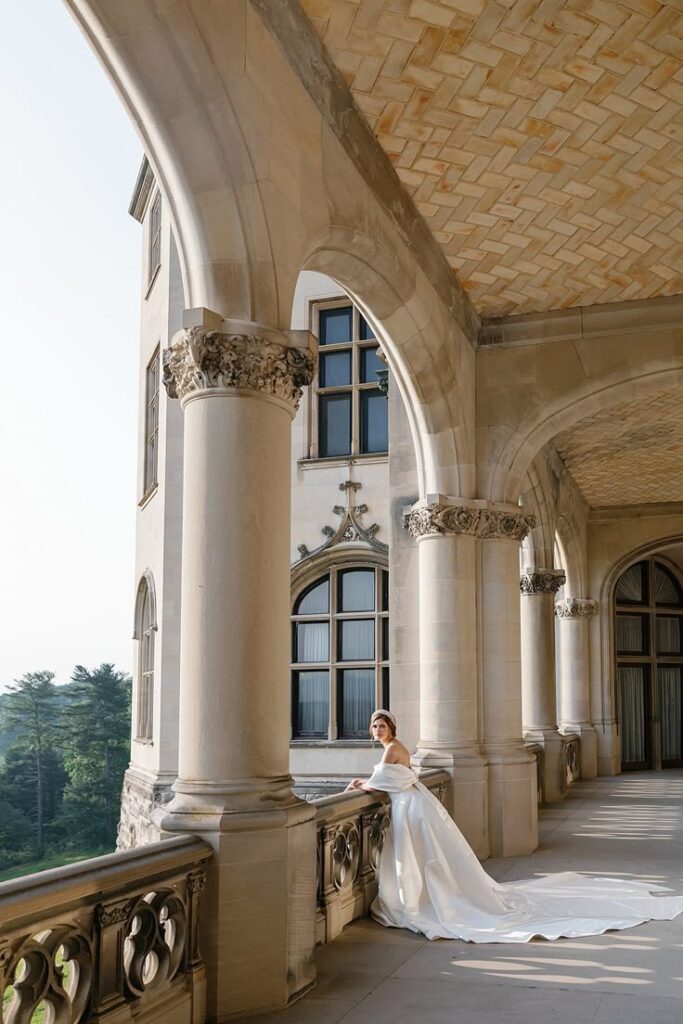 A bride in a classic white gown stands alone on the arched stone balcony of the Biltmore Estate, framed by ornate columns and architectural details.