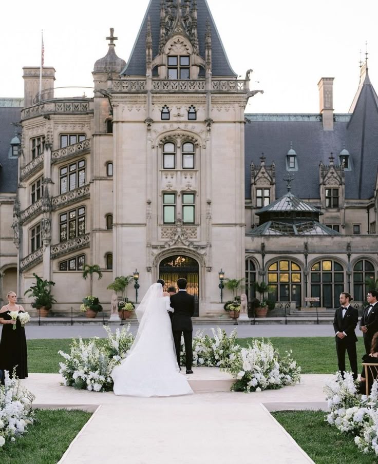 A bride and groom exchange vows in front of the Biltmore Estate, surrounded by white floral arrangements and flanked by bridesmaids and groomsmen in black.