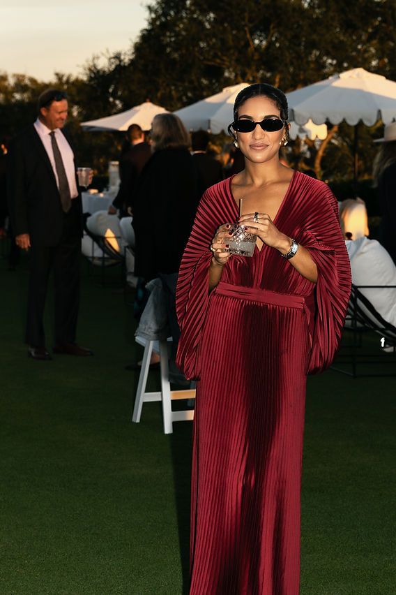Wedding guest in a merlot pleated gown with sculptural sleeves and dark sunglasses, holding a coupe cocktail at an outdoor evening reception — bold, moody fashion for an editorial-style wedding.