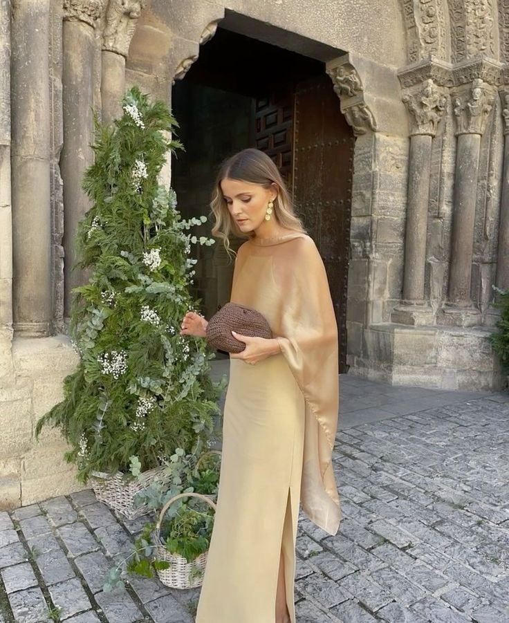 Wedding guest in a gold satin gown and sheer shawl holding a woven clutch, standing outside a historic venue — refined, minimalist wedding guest outfit inspiration.