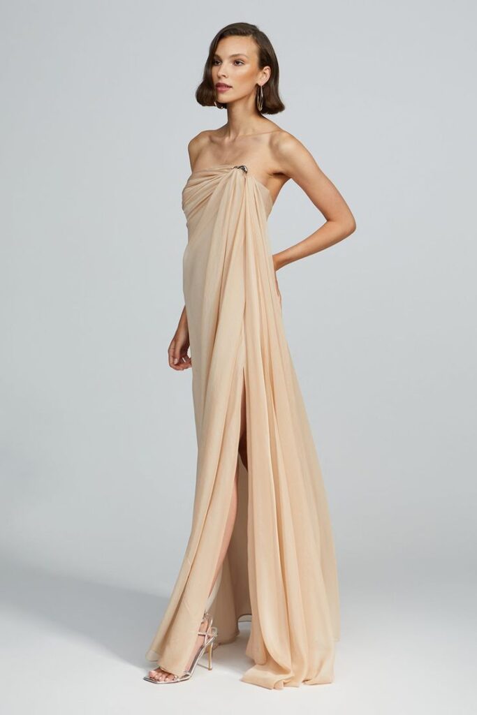 A statuesque woman wears a minimalist, strapless champagne gown with Grecian draping and a high leg slit. Delicate and sculptural, the look evokes coastal cliffs at dusk—light, ethereal, and timelessly modern.