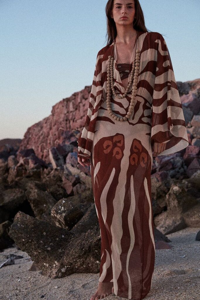 A barefoot woman stands on a rocky shoreline in a maroon and sand patterned caftan, layered with chunky wooden beads. Her gaze is grounded, her presence textural and raw—echoing the natural palette of sun-bleached stone and salt-washed terrain.