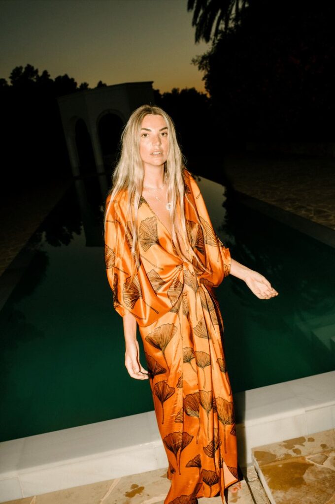 A sunkissed woman stands poolside at golden hour in a fluid, kimono-style gown in burnt orange silk, patterned with abstract ginkgo leaves. Her effortless glow and relaxed silhouette channel vacation elegance with a bold, botanical twist.