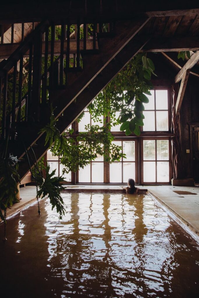 A serene indoor hot spring pool beneath a staircase at Dunton Hot Springs, with a woman relaxing at the water’s edge surrounded by lush greenery and rustic wood beams.