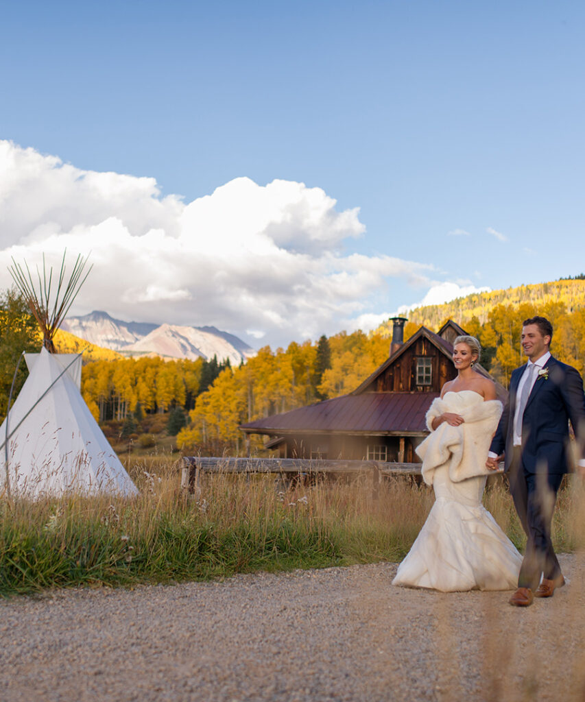 A bride and groom walk hand in hand along a gravel path at Dunton Hot Springs, passing a tipi and a weathered wooden cabin, with golden aspens and mountain peaks in the background.