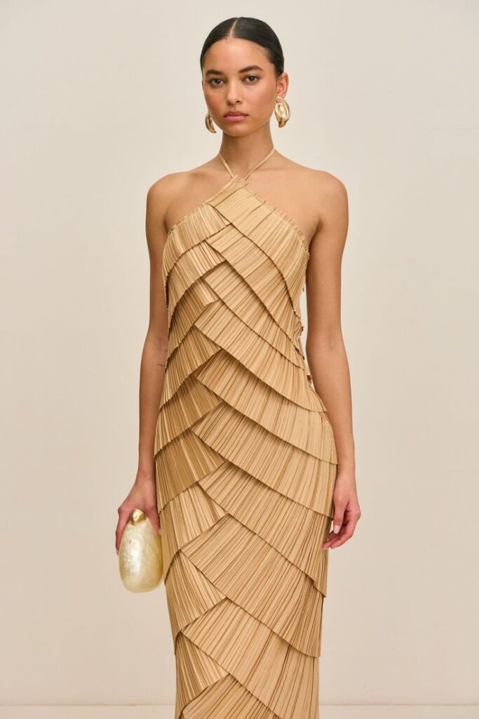 Wedding guest in a sculptural tan halter dress with pleated layers and gold accessories — elevated neutral guest outfit for an earthy and refined wedding.