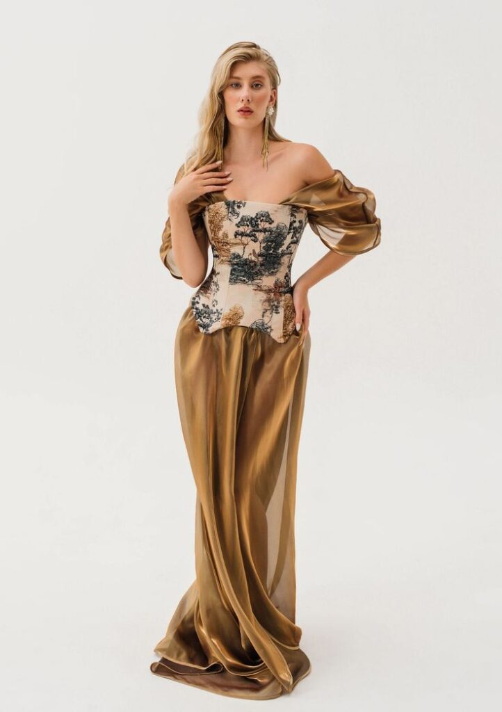 Model in an off-shoulder antique gold gown featuring a structured toile-print corset and sheer organza skirt, evoking regal elegance and heirloom charm.
