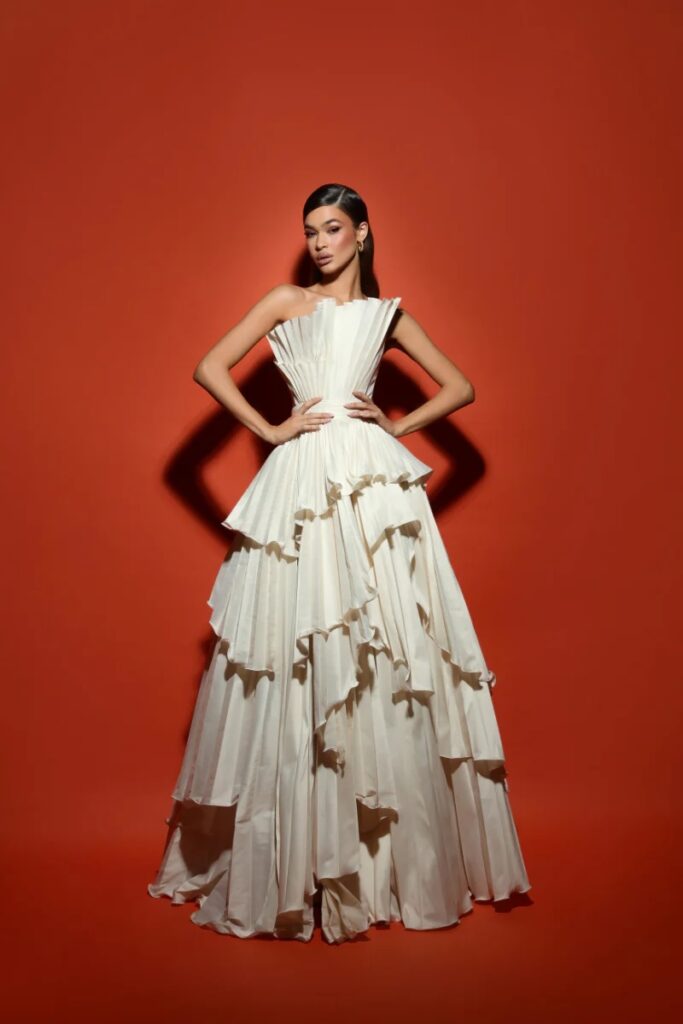 Model in an ivory tiered gown with sculptural pleats and an asymmetrical ruffled silhouette, posed against a rich terracotta background. A modern heirloom moment with regal flair and architectural elegance.