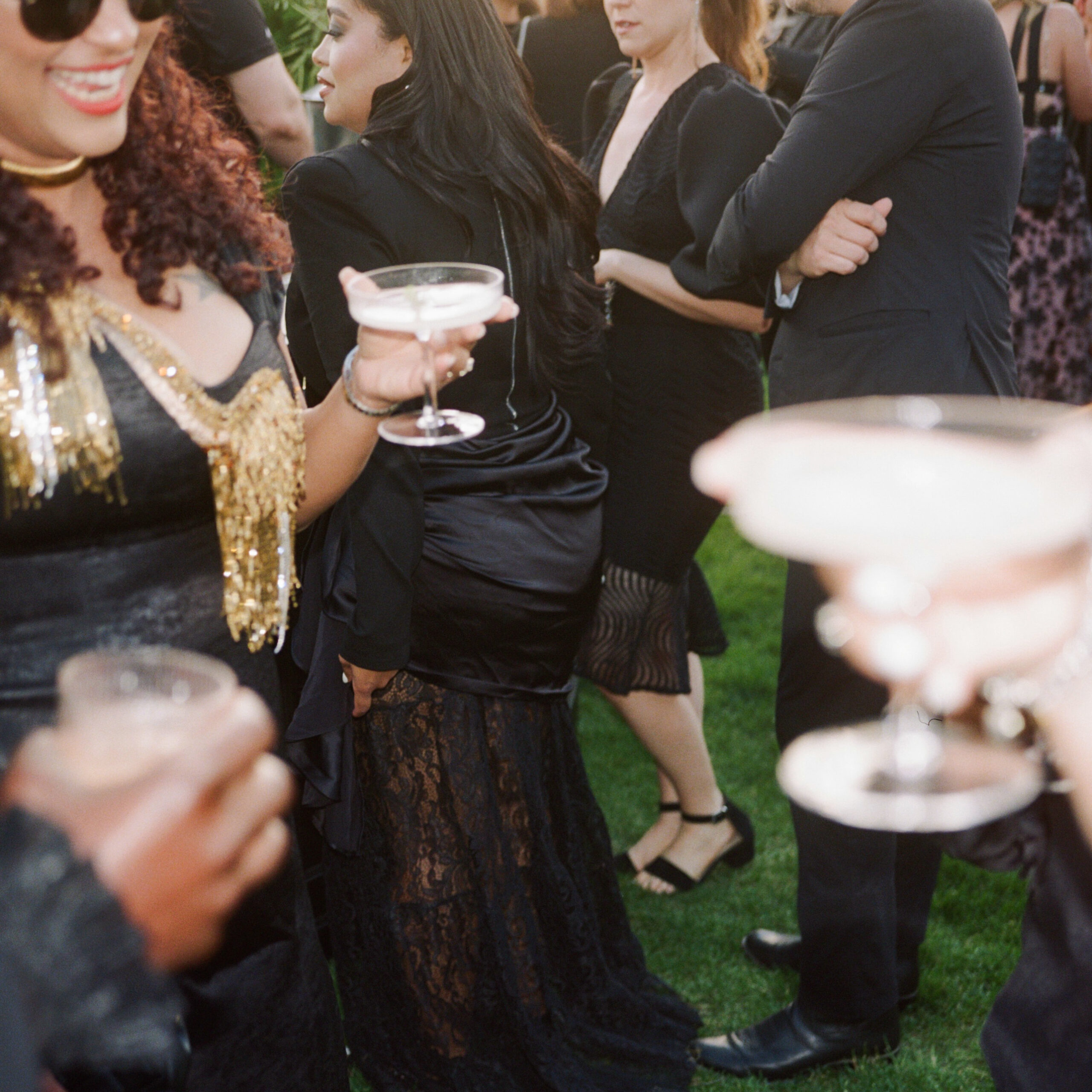 Wedding guests in formal black attire holding coupe cocktails, featuring lace, satin, and gold textures at an outdoor celebration.