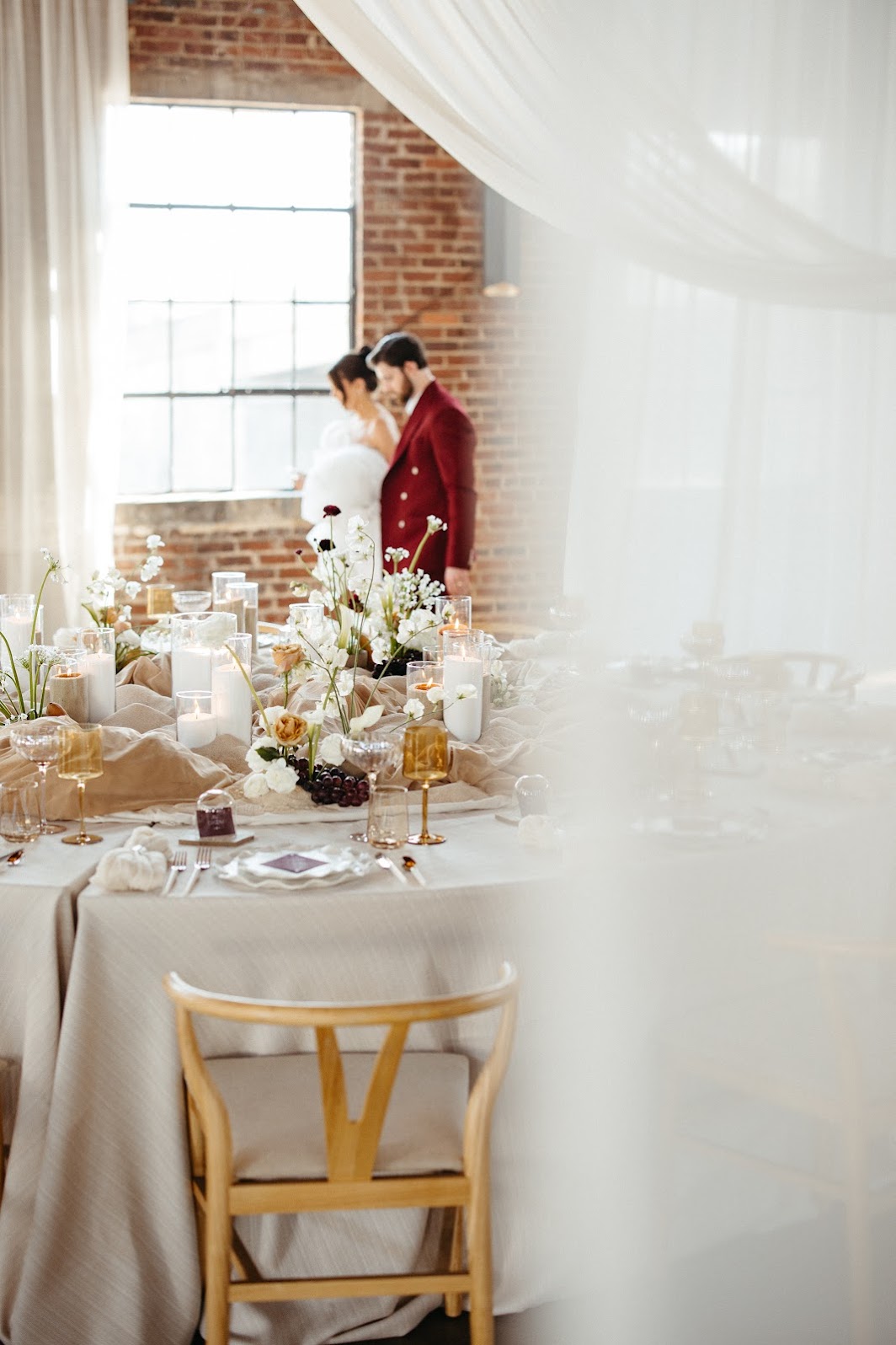 Modern wedding tablescape with neutral draped linens, sculptural florals, and a blurred couple embracing in the background by a sunlit window.