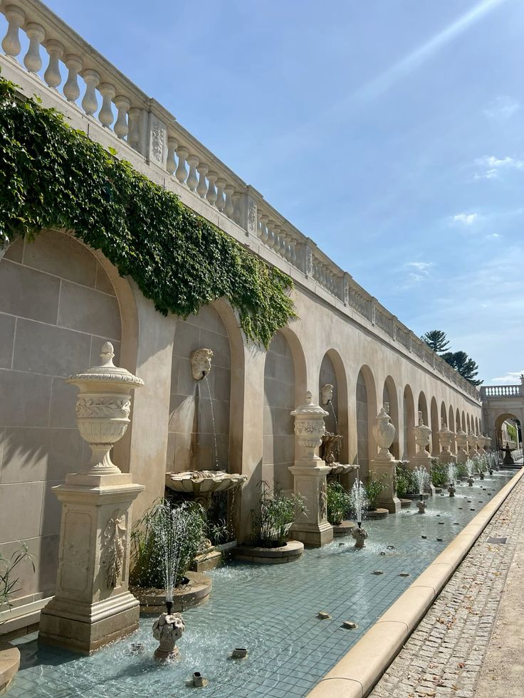Elegant fountain-lined loggia at Longwood Gardens, featuring arched stone walls with sculpted busts, cascading water, and ivy-draped architectural details under a blue sky.