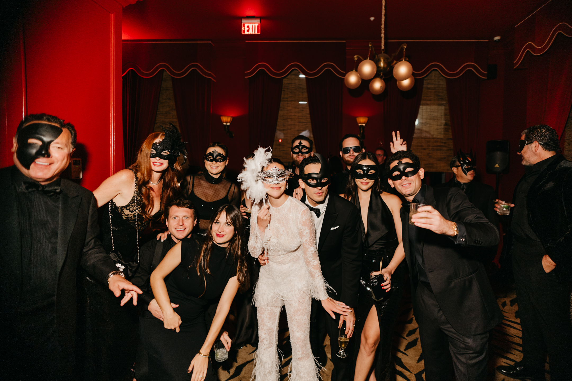 A stylish group of guests in black-tie attire and ornate masks gather around the bride and groom at a lavish masquerade-themed wedding celebration.