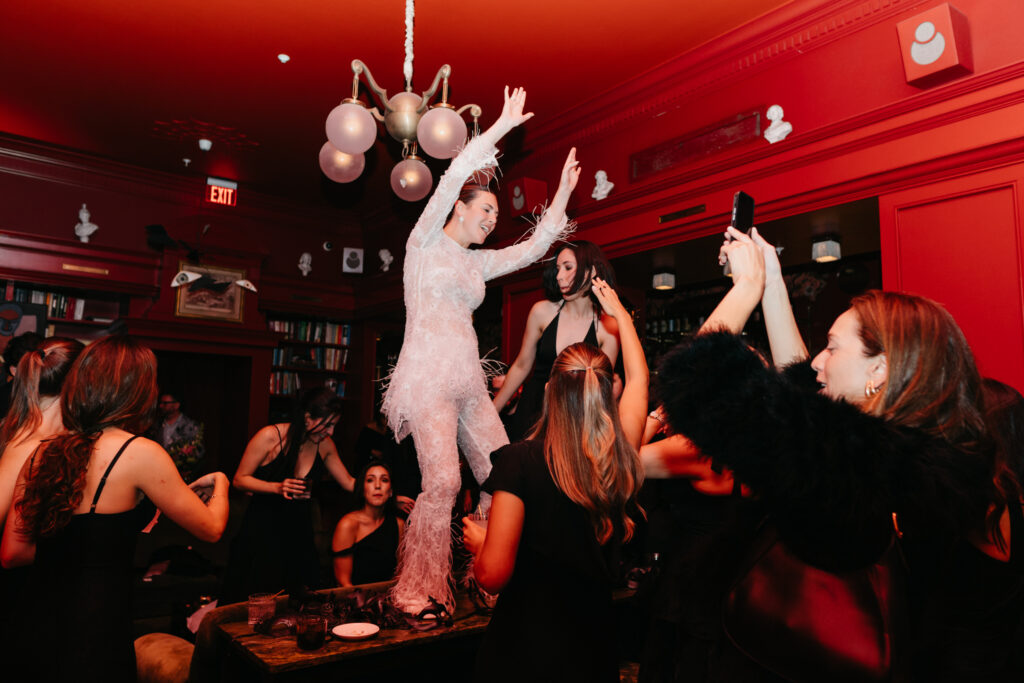 Bride in a feathered jumpsuit dancing on a table surrounded by guests in a red-lit lounge during an energetic wedding after-party.
