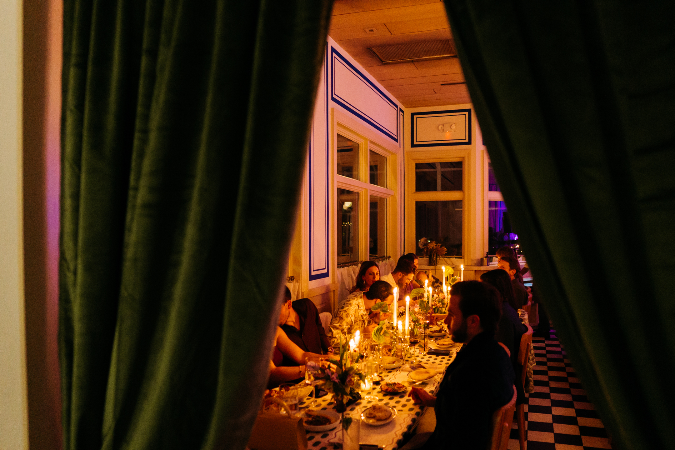 Candlelit dinner scene viewed through emerald velvet curtains, with guests seated around a long table set with patterned linens, taper candles, and layered place settings.